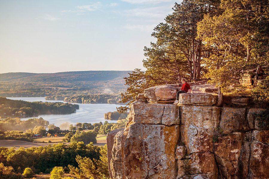 Contact - Aerial View of Trees, River and Rock Ledge in Wisconsin With a Person Sitting Close to Edge of a Cliff at Dusk